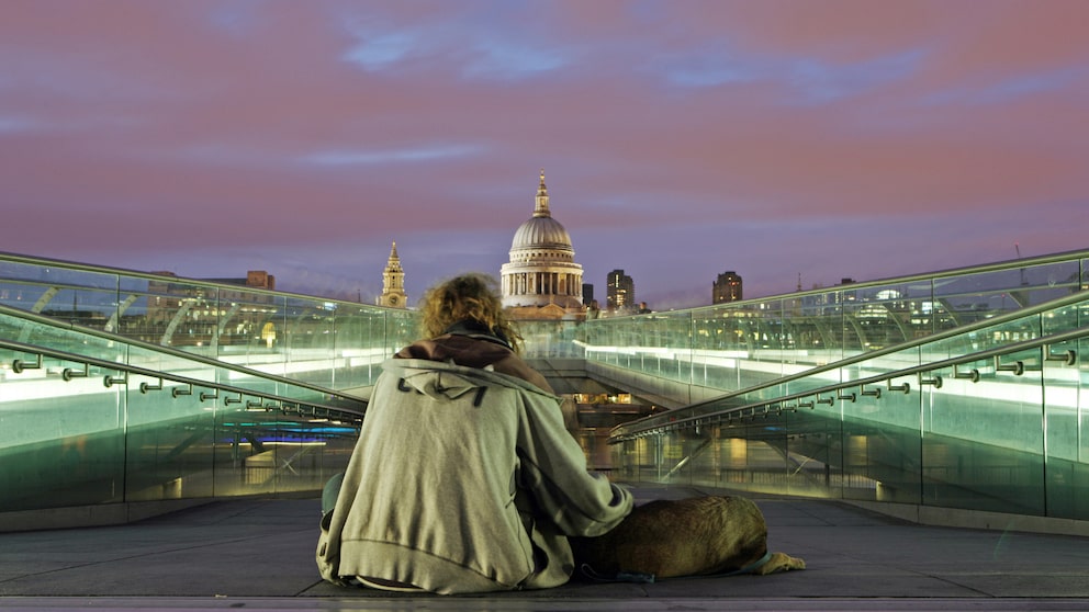 Ein obdachloser Mann und sein Hund bei Nacht auf der Millennium Bridge mit der St. Paul's Cathedral im Hintergrund.