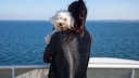 Frau mit weißem Hund auf dem Arm steht auf dem Deck einer Fähre und schaut aufs Meer