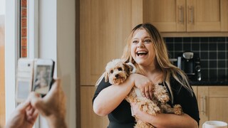 Hund hält ihren Pudel wie ein Baby im Arm. Person mit Handy filt sie dabei