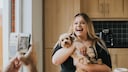 Hund h&auml;lt ihren Pudel wie ein Baby im Arm. Person mit Handy filt sie dabei