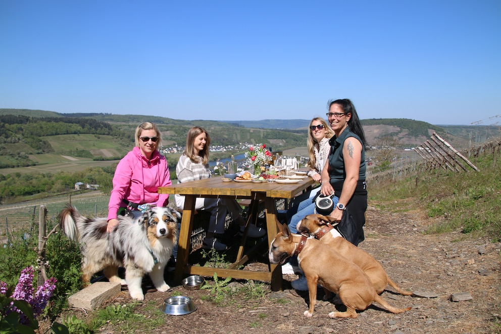 Vier Frauen sitzen mit drei Hunden an einem gedeckten Tisch im Freien.