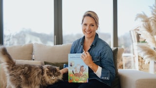 Judith Rakers sitzt mit einer Katze auf einer Couch und präsentiert ihr Kinderbuch.