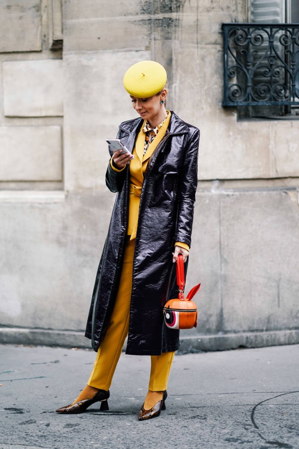 PARIS, FRANCE - MARCH 01: A guest wears a yellow beret hat, a yellow pantsuit, earrings, a shiny black vinyl raincoat, an orange-shaped handbag, a shiny golden python pattern pointy pumps with fantasy heels, yellow socks , outside Issey Miyake, during Paris Fashion Week Womenswear Fall/Winter 2019/2020, on March 01, 2019 in Paris, France. (Photo by Edward Berthelot/Getty Images)
