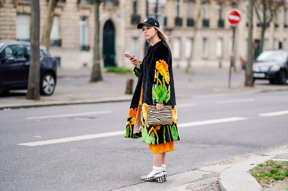 PARIS, FRANCE - FEBRUARY 28: A guest wears a NY black cap, a black fluffy coat with yellow,orange and green daffodils design, an orange dress, a yellow and black zebra pattern bag, white openwork socks, deep green and white gingham slingback pumps, outside Atlein, during Paris Fashion Week Womenswear Fall/Winter 2019/2020, on February 28, 2019 in Paris, France. (Photo by Edward Berthelot/Getty Images)