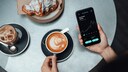 Frauenhände, mit Handy, auf dem Aktienkurse verzeichnet sind. Vor ihr Kaffee und Croissant.