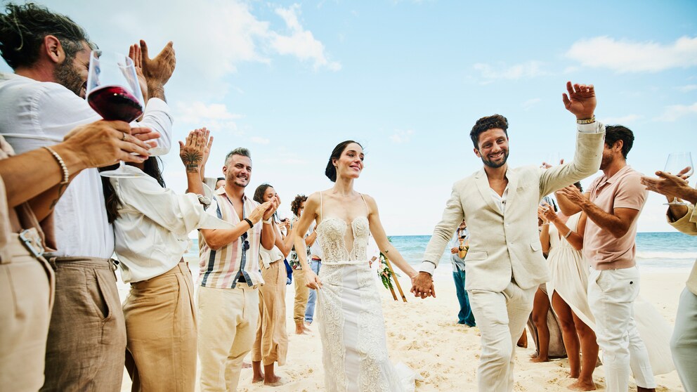 Er und sie in Weiß am Strand umgeben von klatschenden Leuten in Leinen-Weiß. Hach, Strand – ob mit oder ohne Hochzeit schön, oder?