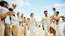 Er und sie in Weiß am Strand umgeben von klatschenden Leuten in Leinen-Weiß. Hach, Strand – ob mit oder ohne Hochzeit schön, oder?