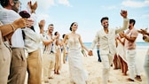 Er und sie in Weiß am Strand umgeben von klatschenden Leuten in Leinen-Weiß. Hach, Strand – ob mit oder ohne Hochzeit schön, oder?