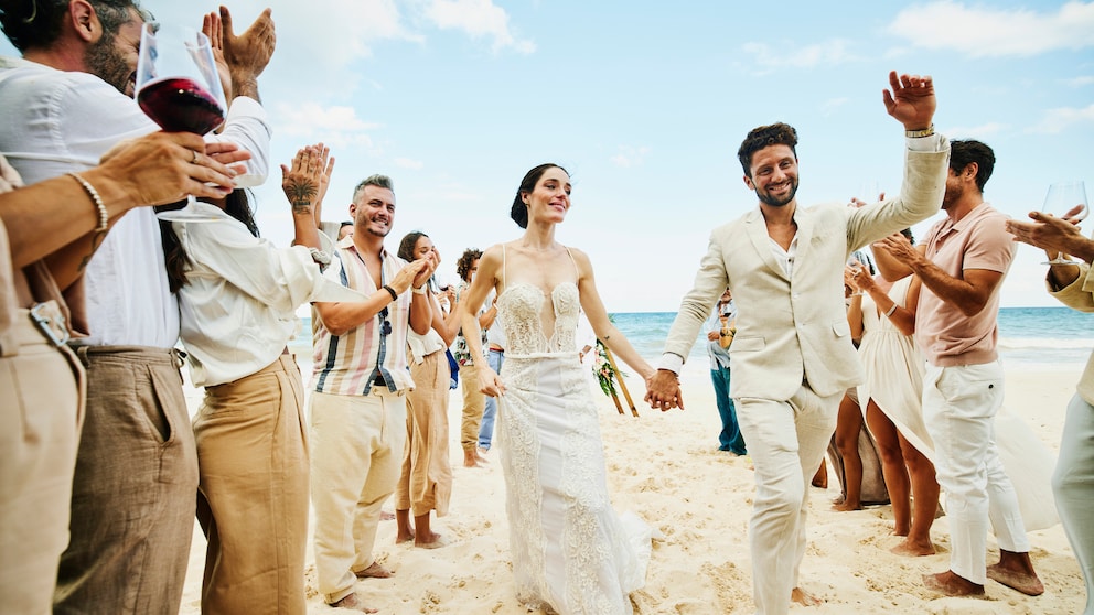 Er und sie in Weiß am Strand umgeben von klatschenden Leuten in Leinen-Weiß. Hach, Strand – ob mit oder ohne Hochzeit schön, oder?