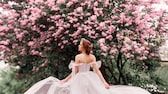 Braut in rosé-farbenem Off-Shoulder-Kleid vor einem blühenden Kirschbaum. Sie blickt zum Baum, das leichte Kleid weht, wunderschön.