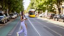 Junge Frau in weißem Top und fliederfarbener weiter Stoffhose überquert lachend Tramschienen