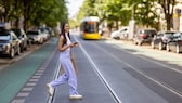 Junge Frau in weißem Top und fliederfarbener weiter Stoffhose überquert lachend Tramschienen