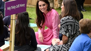 Prinzessin Kate im pinken Kleid. Sie sitzt auf dem Boden neben einigen Kindern, denen sie Rede und Antwort steht und grinst