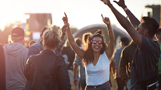 Junge Frau mit weißem Top und Shorts umgeben von Freundinnen feiert mit den Händen in der Luft und lacht. Sie trägt einen Messy Bun und eine Sonnenbrille und sieht wirklich so aus als hätte sie Spaß, ohne gestellt zu wirken