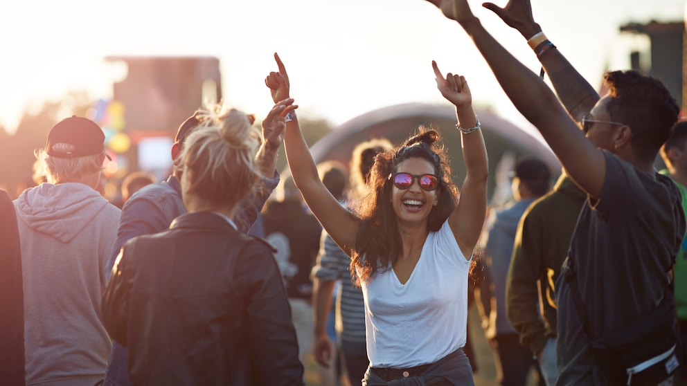 Junge Frau mit weißem Top und Shorts umgeben von Freundinnen feiert mit den Händen in der Luft und lacht. Sie trägt einen Messy Bun und eine Sonnenbrille und sieht wirklich so aus als hätte sie Spaß, ohne gestellt zu wirken