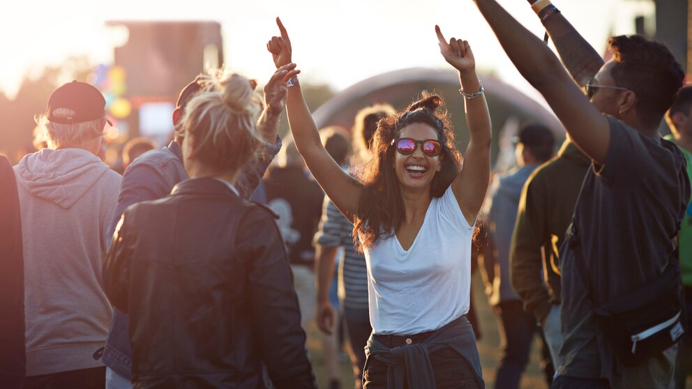 Junge Frau mit weißem Top und Shorts umgeben von Freundinnen feiert mit den Händen in der Luft und lacht. Sie trägt einen Messy Bun und eine Sonnenbrille und sieht wirklich so aus als hätte sie Spaß, ohne gestellt zu wirken