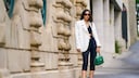 Frau in den Straßen von Paris in dunklen, engen Caprihosen mit schwarzem Top, weißem Blazer und grüner Handtasche