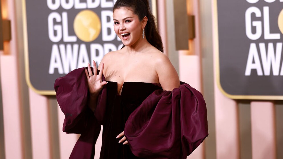 Selena Gomez im schwarzen Off-Shoulder-Kleid mit auberginefarbener Scherpe wie sie lachend in die Kamera winkt