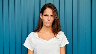 Junge Frau im weißen T-Shirt vor blauer Wand mit grantigem Gesicht