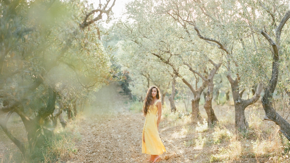 Junge Frau im gelben Sommerkleid mit offenen Haaren zwischen Olivenbäumen. Die Sonne fällt durch die Bäume, es wirkt alles malerisch und verträumt und passt damit, wie ich finde, ideal zur „Tomato Girl“-Ästhetik.