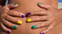 Frau mit bunt lackierten Nägeln hält sich die Hande auf den gebräunten Babybauch