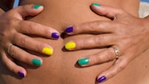 Frau mit bunt lackierten Nägeln hält sich die Hande auf den gebräunten Babybauch