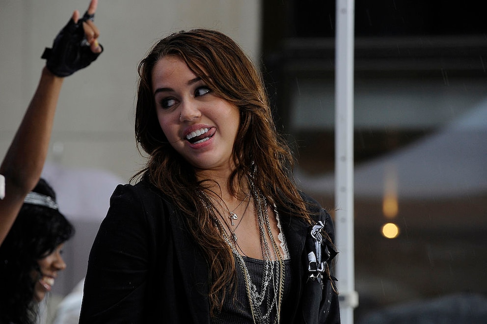 Miley Cyrus mit brünetten Haaren bei einem Auftritt in New York im August 2009. Sie trägt viele KEtten, die Haare sehen chaotisch aus, bisschen als wäre sie nass geworden. Die damals 16-Jährige streckt die Zunge raus