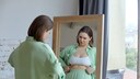 Frau in grünem Ensemble mit weißem Crop-Top vor dem Spiegel. Prüfend kneift sie sich in den Bauch