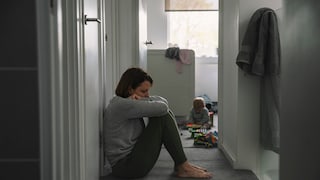 Junge Frau sitzt im Flur auf dem Boden, den Kopf auf die Knie gestützt. Im Hintergrund spielt ein Baby