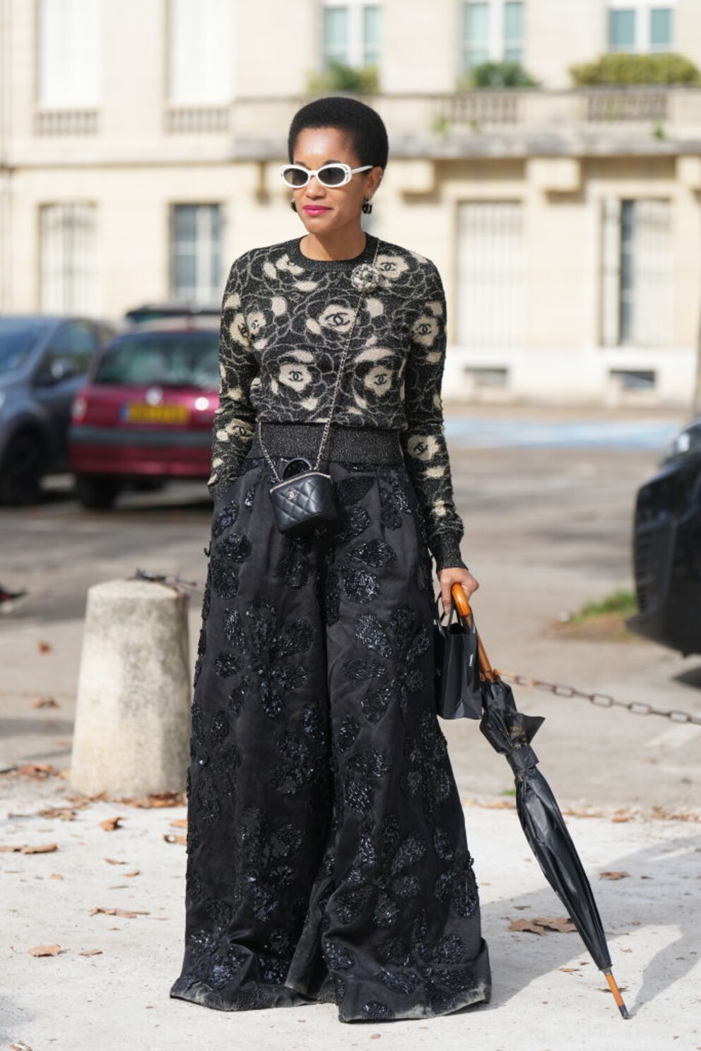 Junge Frau in Paris mit Chanel-Blumen-Strickpulli und schwarzem Maxirock und Regenschirm. Sehr stimmiges Outfit