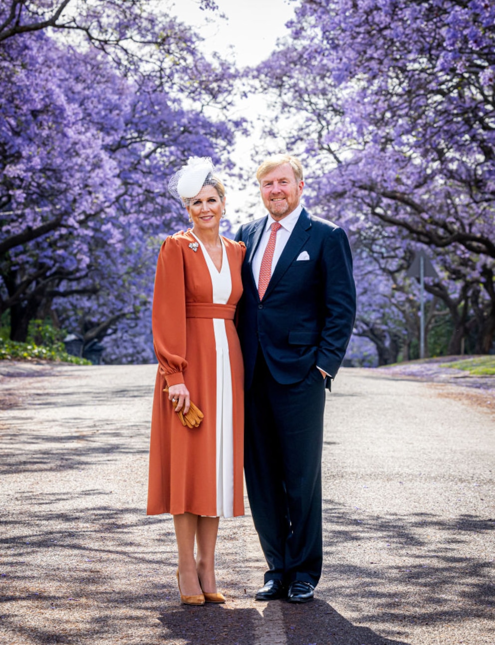 Königin Máxima und König Willem-Alexander vor einem Meer aus fliederfarbenen Blüten. Sie trägt ein orange-weißes Kleid, er Basic-Anzug