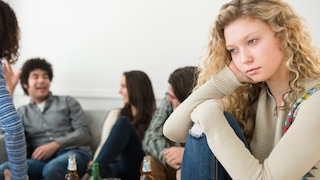 Frau mit blonden Locken sitzt abseits einer Gruppe, die sich angeregt unterhält und guckt in die Ferne
