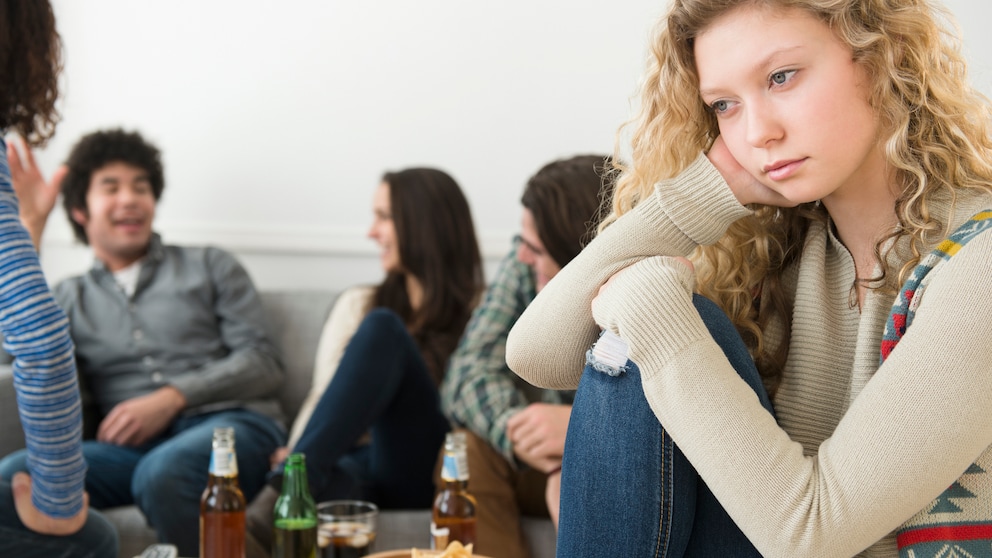 Frau mit blonden Locken sitzt abseits einer Gruppe, die sich angeregt unterhält und guckt in die Ferne