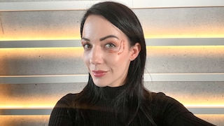 Carmen mit Hot Glue im Gesicht, wie sie versucht, freundlich in die Kamera zu gucken, ohne dass ihr Auge hängt wegen des schweren Heißklebedesigns und so, dass man eben dieses noch gut erkennen kann
