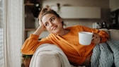 Frau im orangen Sweatshirt auf dem Sofa mit einer Tasse Tee