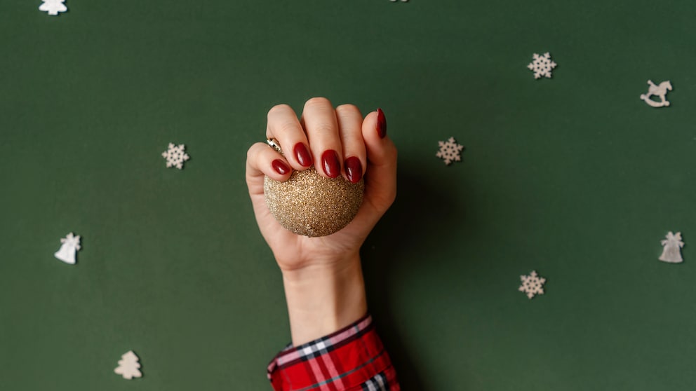 Hand mit rot lackierten Nägeln, die eine gold glitzernde Baumkugel hält vor grünem Hintergrund