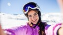 Frau mit pinker Schneejacke und Skibrille, die ein Selfie zu machen scheint vor Schneelandschaft