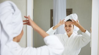 Frau massiert die Stirn vor dem Spiegel in weißem Bademantel und mit Handtuch auf dem Kopf. Ein weniger gestelltes Foto wäre mir lieber gewesen, aber es ist schwer, was passendes zur Gesichtsmassage zu finden