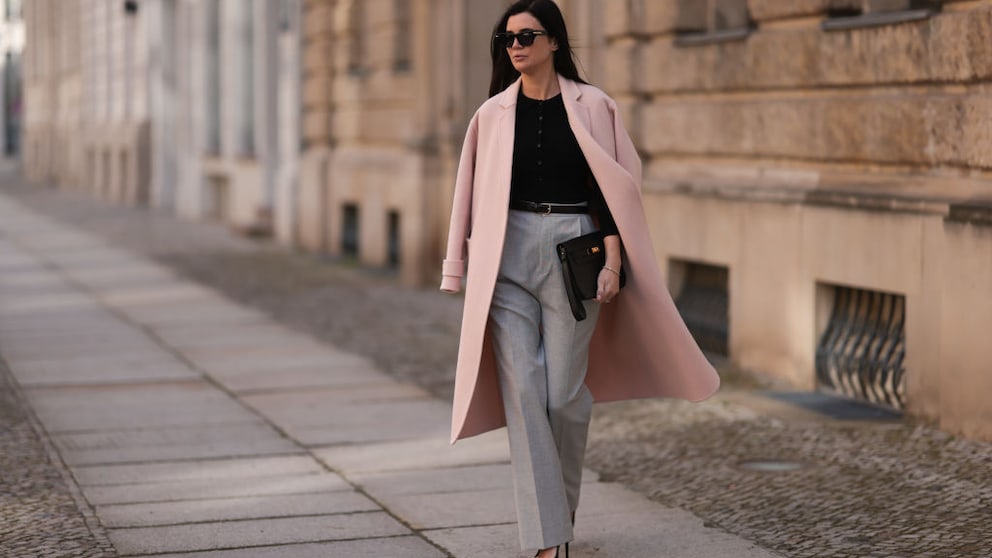 Frau mit Anzughose in Grau, schwarzem Oberteil und Sonnenbrille, dazu rosa Mantel. Streetstyle