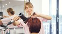 Blonde Friseurin föhnt Kundin mit roter Kurzhaarfrisur die Haare, Gesicht zur Kamera gewandt und leicht lächelnd