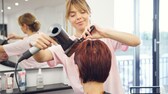 Blonde Friseurin föhnt Kundin mit roter Kurzhaarfrisur die Haare, Gesicht zur Kamera gewandt und leicht lächelnd