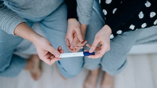 Overhead view of an affectionate young Asian couple sitting on the bed, holding hands and holding a positive pregnancy test together. It's finally happening. The long-awaited news. Life events, fertility and family concept