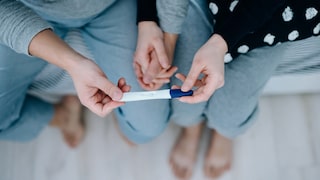 Overhead view of an affectionate young Asian couple sitting on the bed, holding hands and holding a positive pregnancy test together. It's finally happening. The long-awaited news. Life events, fertility and family concept