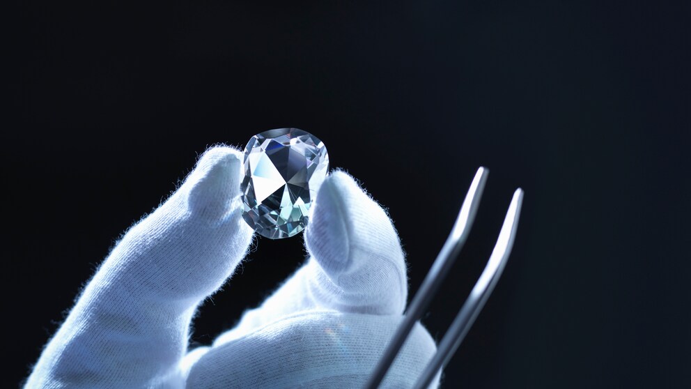 Behandschuhte Hand hält Diamant und Zange vor schwarzem Hintergrund