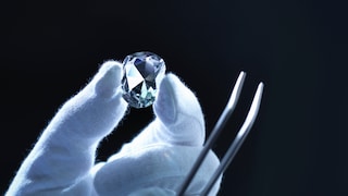 Behandschuhte Hand hält Diamant und Zange vor schwarzem Hintergrund