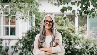 Frau mit langen Haaren, grau am Ansatz und Längen, Spitzen noch dunkel vor hübschem Haus-Garten-Setting