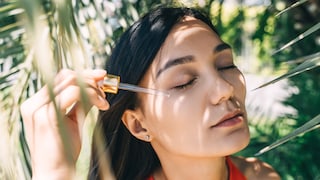 Frau, die sich mit Pipette etwas Pflegeprodukt ins Gesicht träufelt mit geschlossenen Augen vor einem Palmenhintergrund im orangen Top