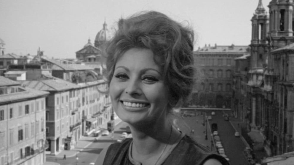 Sophia Loren lächelt sehr süß in die Kamera. Hinter ihr eine italienische Stadtkulisse