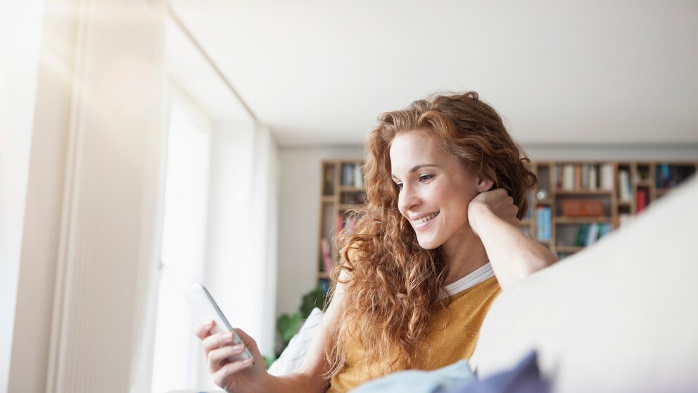 Frau grinst ins Handy auf dem Sofa. Es sei ihr zu wünschen, dass sie eine süße Nachricht bekommen hätte, anstatt online zu shoppen