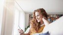 Frau grinst ins Handy auf dem Sofa. Es sei ihr zu wünschen, dass sie eine süße Nachricht bekommen hätte, anstatt online zu shoppen